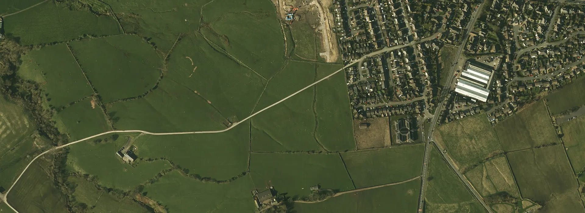 An aerial view of a rural landscape showing green fields divided by hedgerows and paths, with a residential area on the right side.