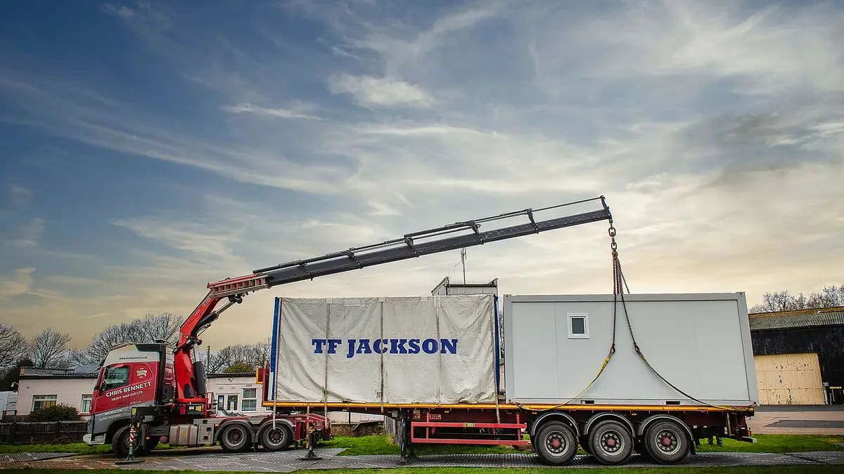A red crane truck lifting a modular building unit with 'TF Jackson' branding, set against a blue sky