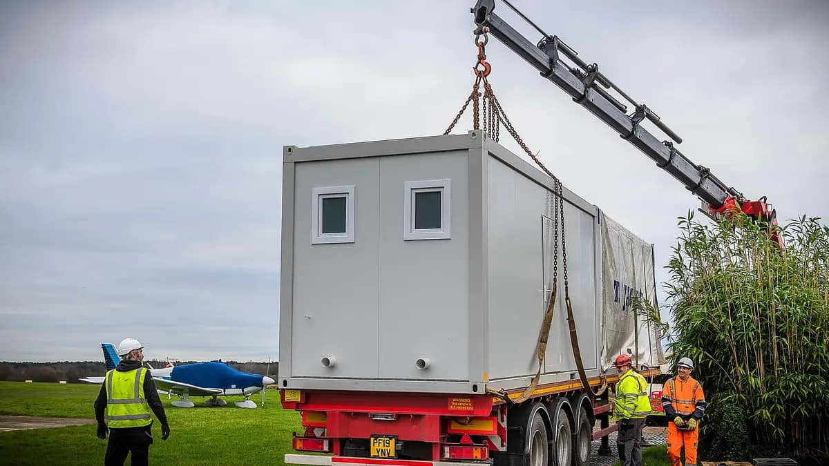 Modular building unit being lifted by a crane onto a truck, with workers supervising, near an airfield