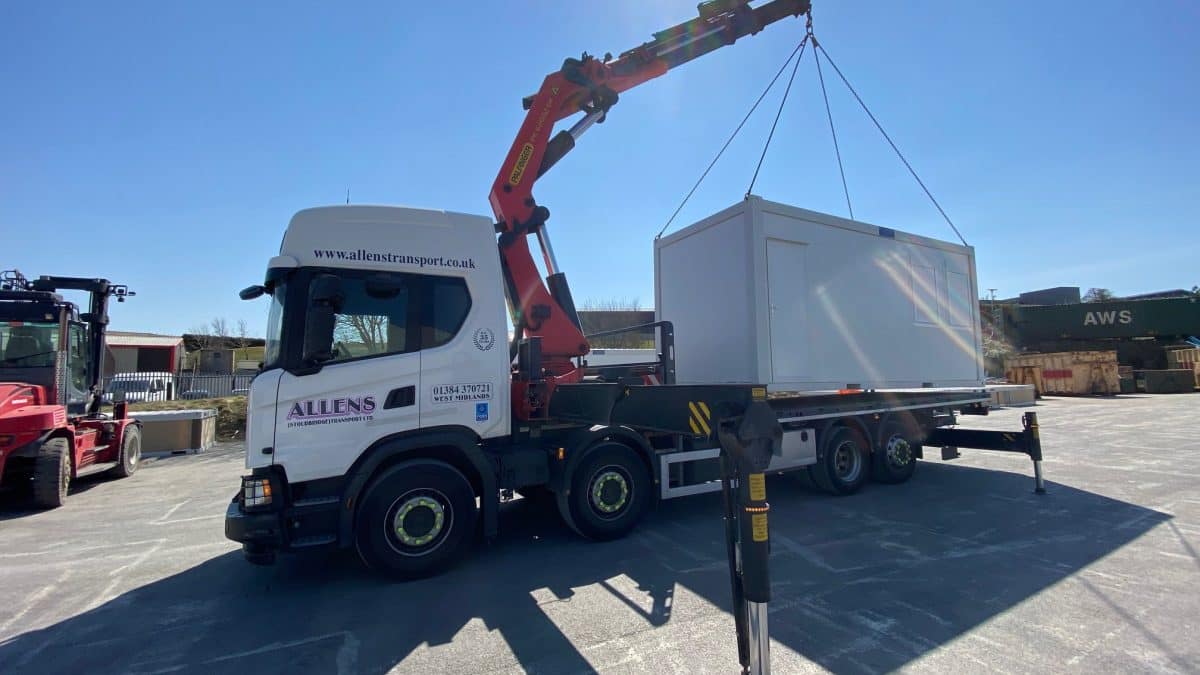 A truck with a crane lifting a modular unit in an industrial area under a clear sky.