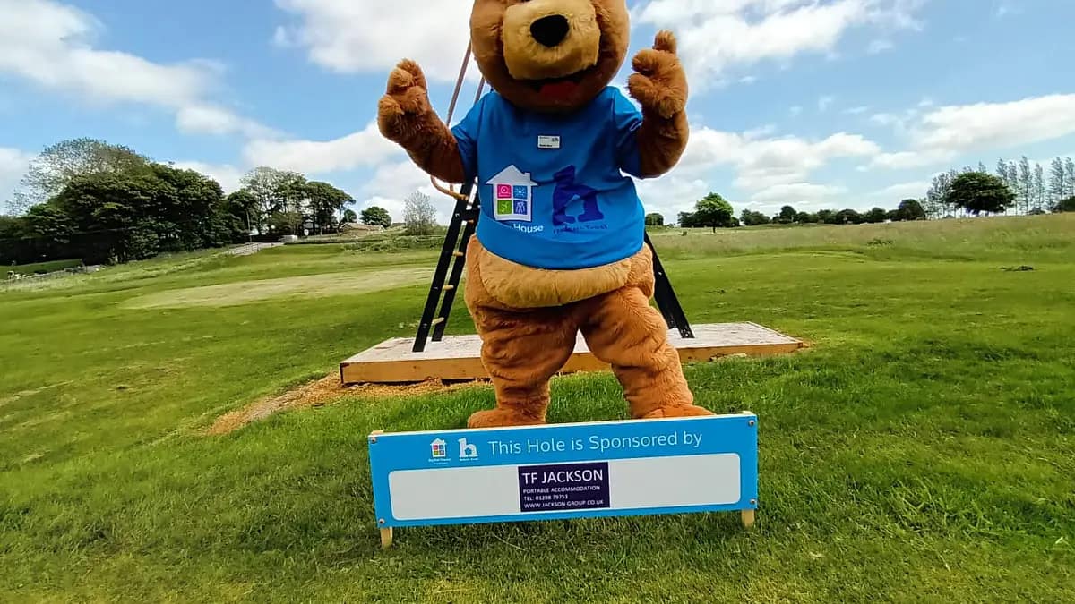 mascot bear in a blue t-shirt waving above a sign stating 'This Hole is Sponsored by TF JACKSON,' on a sunny golf course