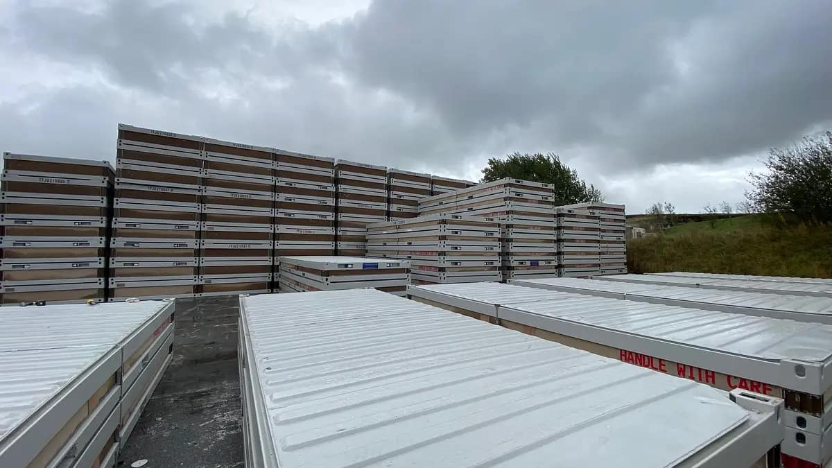 Stacked portable metal containers beside flatbed trailers with 'Handle with Care' signage under an overcast sky.