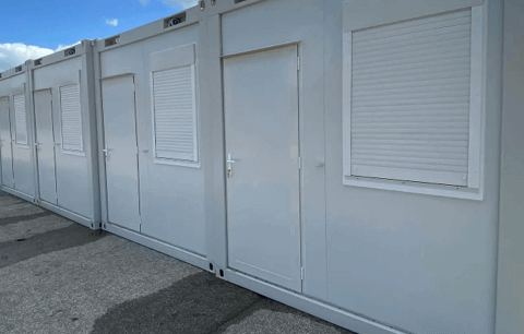 Row of portable sleeping units with white shutters in an outdoor setting, under a clear sky