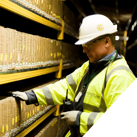 A professional wearing a hard hat and reflective jacket examines a shelf filled with labeled archive boxes in a high-security storage area.
