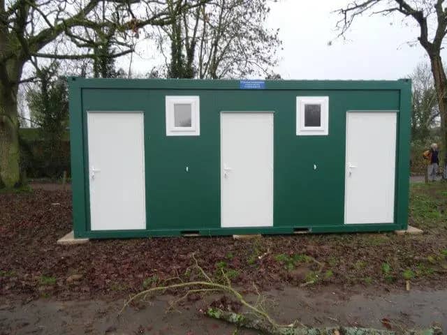 Green modular building with three doors and windows in a leafy setting.