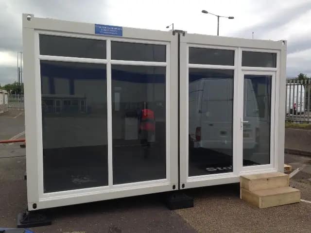 Exterior of a modular office with large glass doors and a reflection of a man and a van.