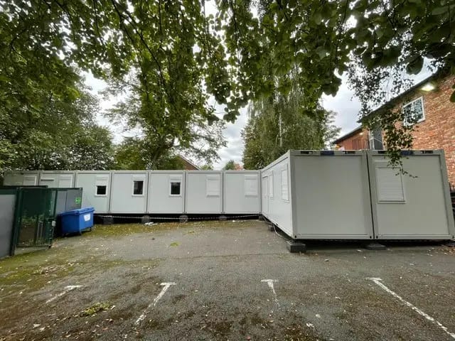 Row of gray modular buildings in a parking area surrounded by trees, with a residential building in the background