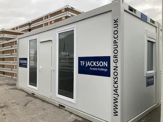 Portable modular building by TF Jackson with double doors, large windows, and company branding, set against a backdrop of stacked modules.