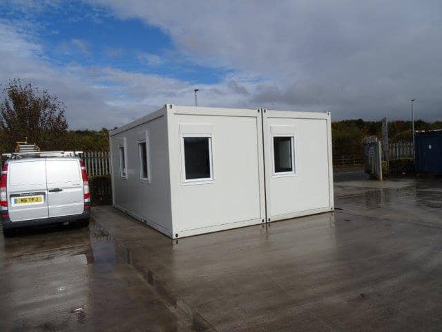 White modular building with two attached sections, set on a wet concrete lot next to a white van