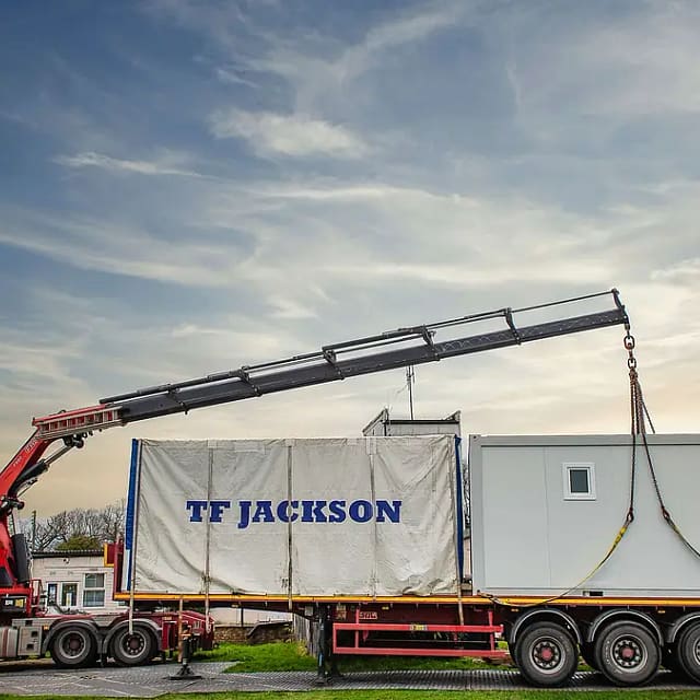 A red crane truck lifting a modular building unit with 'TF Jackson' branding, set against a blue sky