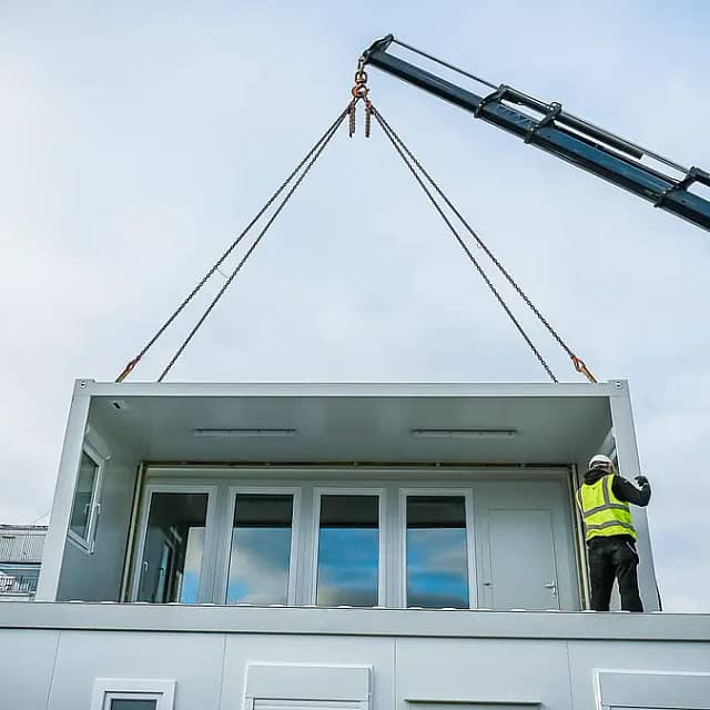 A crane lifting a prefabricated modular section of a building with a worker in high-visibility clothing guiding it into place against a cloudy sky backdrop.