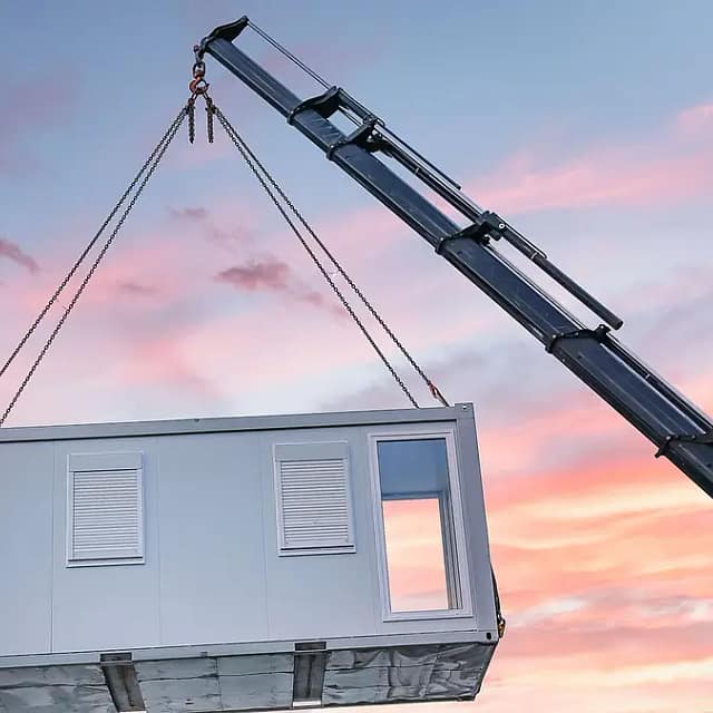 "Alt: A crane lifting a modular building unit against a sunset sky with pink and blue hues