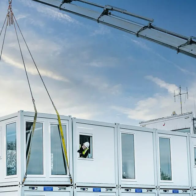 Crane hoisting a modular building unit with a worker assisting, against a blue sky with wispy clouds.
