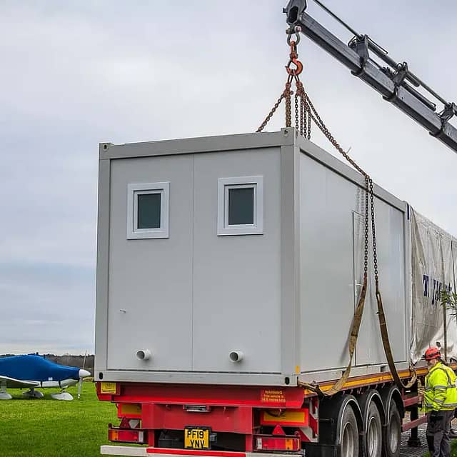 Modular building unit being lifted by a crane onto a truck, with workers supervising, near an airfield