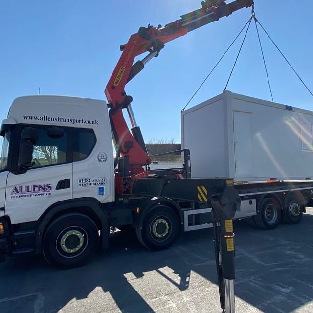 A truck with a crane lifting a modular unit in an industrial area under a clear sky.