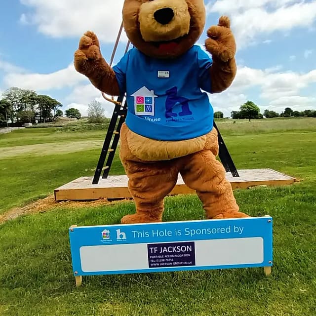 mascot bear in a blue t-shirt waving above a sign stating 'This Hole is Sponsored by TF JACKSON,' on a sunny golf course