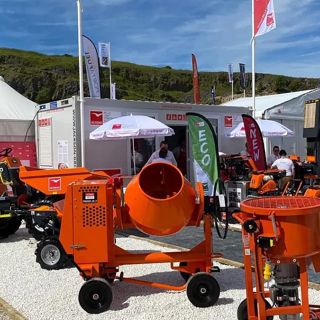 An outdoor exhibition of construction machinery featuring orange mixers and compact tractors, with visitors and tents in the background