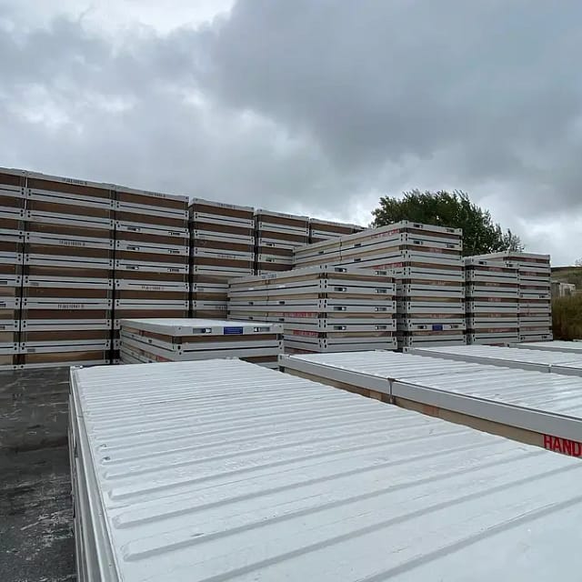 Stacked portable metal containers beside flatbed trailers with 'Handle with Care' signage under an overcast sky.