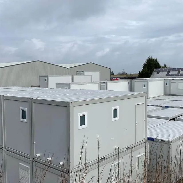 Array of grey modular portable buildings with industrial warehouses in the background, under a cloudy sky