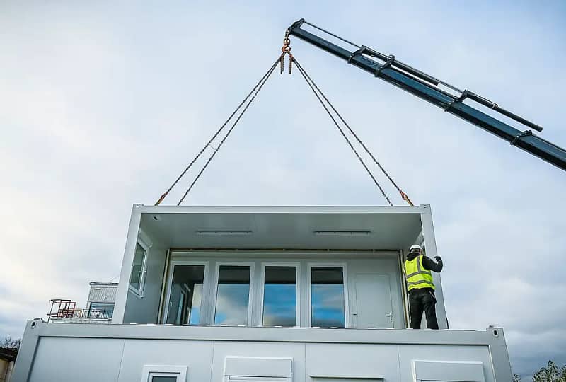 A crane lifting a prefabricated modular section of a building with a worker in high-visibility clothing guiding it into place against a cloudy sky backdrop.