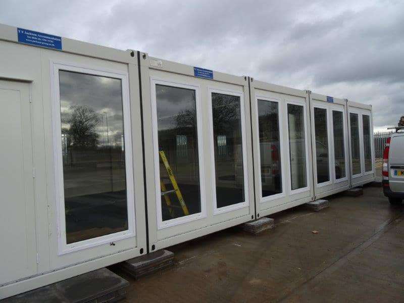 A series of large windows on a modular building, possibly a portable office, with reflections of a cloudy sky