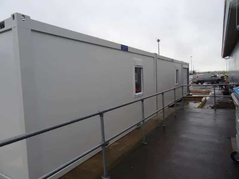 Exterior of a long modular building equipped with small windows, situated on a wet ground next to a railing