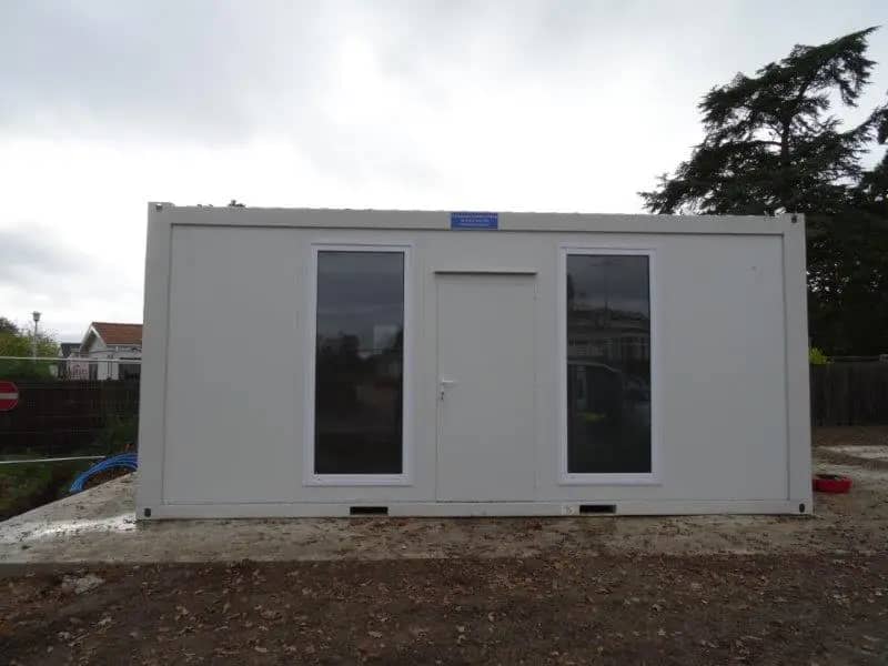 Exterior of a white modular building with glass doors and a flat roof, set on a muddy construction site