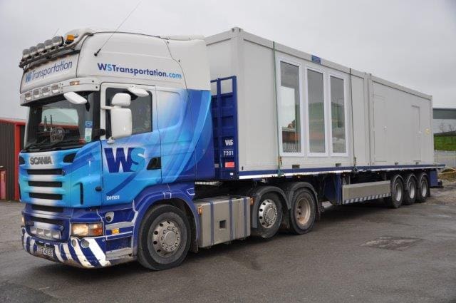 Large truck transporting a white modular building unit on its trailer in a parking lot